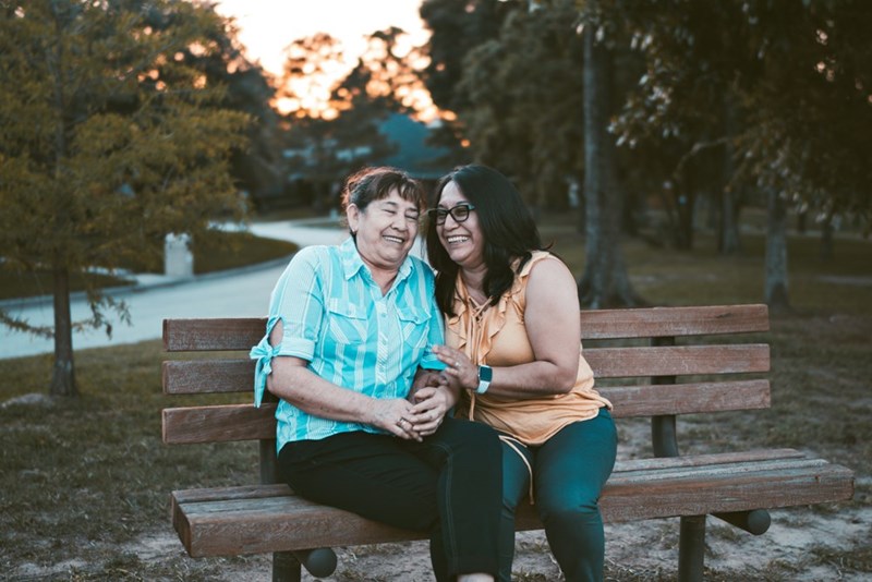 Mother and adult daughter sitting together at a bench in a park. Representation.