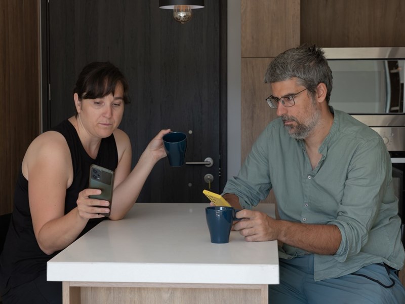 Two adults sit at a kitchen island holding phones and mugs in a modern apartment.