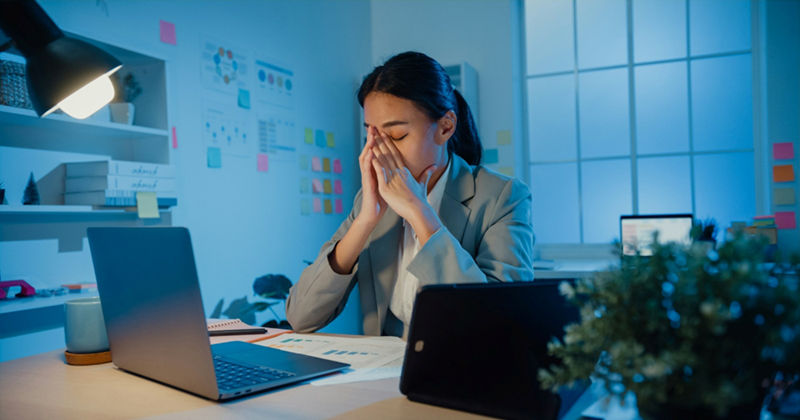 A side view of a model representing an employee sitting at her desk in the evening, touching her hands to her face in frustration
