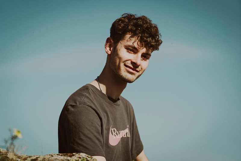 Young man with curly hair smiles slightly outdoors against a clear blue sky.