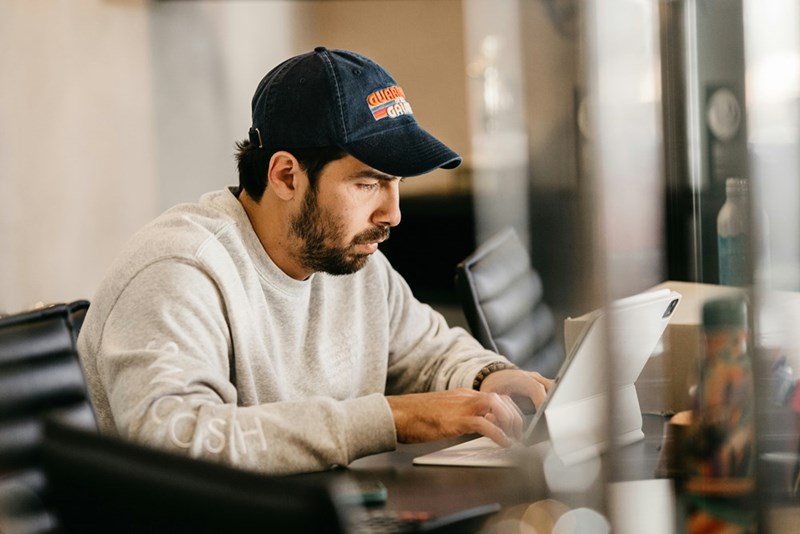 Man in a black cap sits at a counter using a tablet, looking focused indoors.