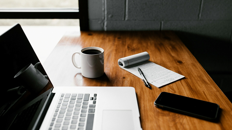 A notebook and pen, phone, laptop, and cup of black coffee on a wooden table