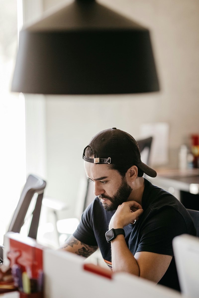 Man in a black cap sits at a desk looking down thoughtfully under a large hanging lamp.
