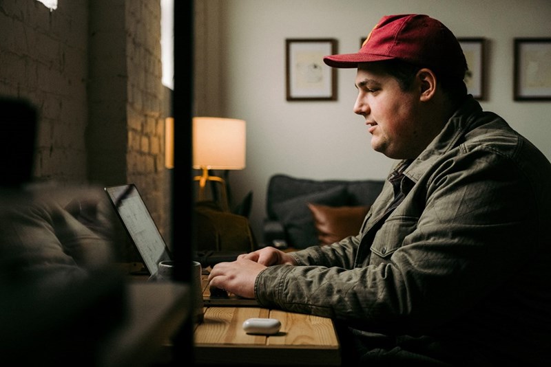 Man in a red cap works on a laptop at a wooden table in a dim room.
