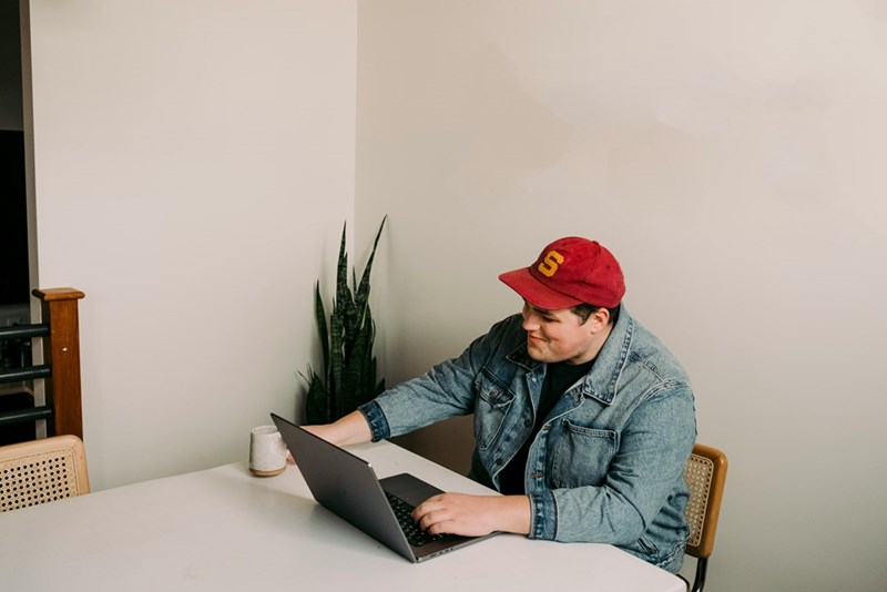 Man in a red cap and denim jacket smiles while using a laptop at a white table.