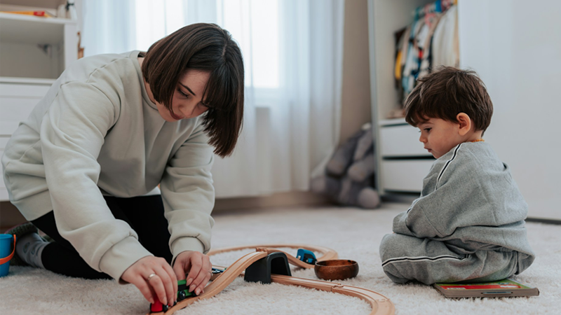 A representation of a woman and a toddler playing with a wooden train set on the floor
