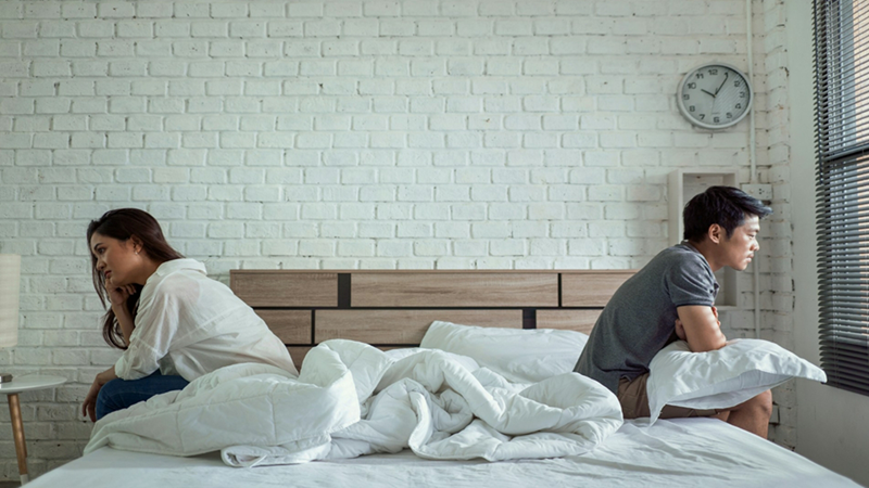 A woman and a man sitting on opposite sides of an unmade bed against a white brick wall