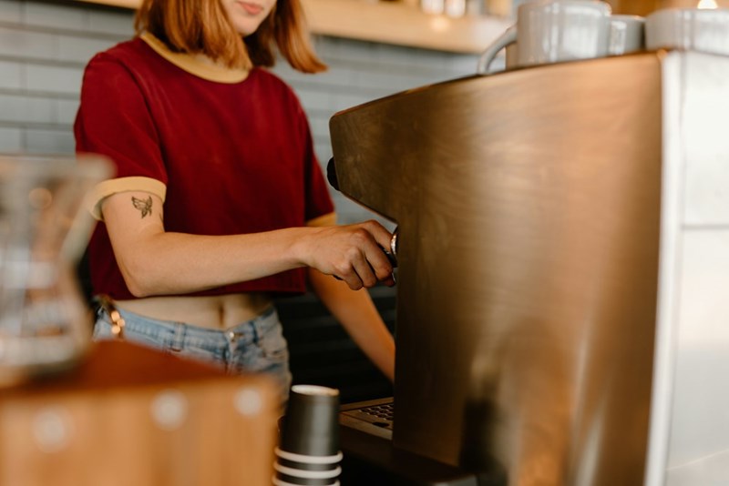 Woman in a red shirt works behind an espresso machine at a cafe counter.