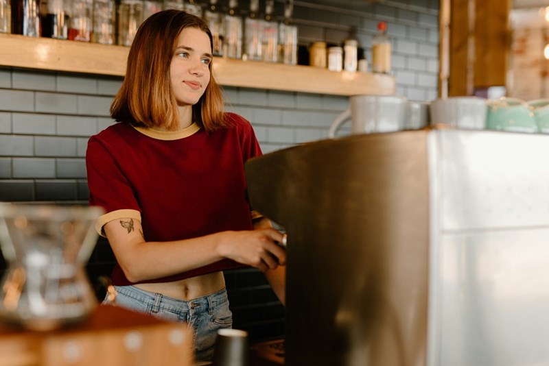 Woman in a red shirt stands behind an espresso machine at a cafe counter.