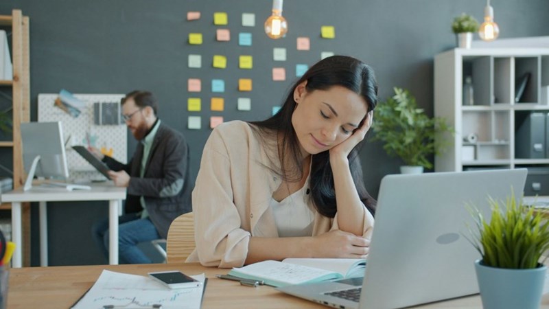 A woman looking sleepy at her computer while another coworker works behind her. Representation.