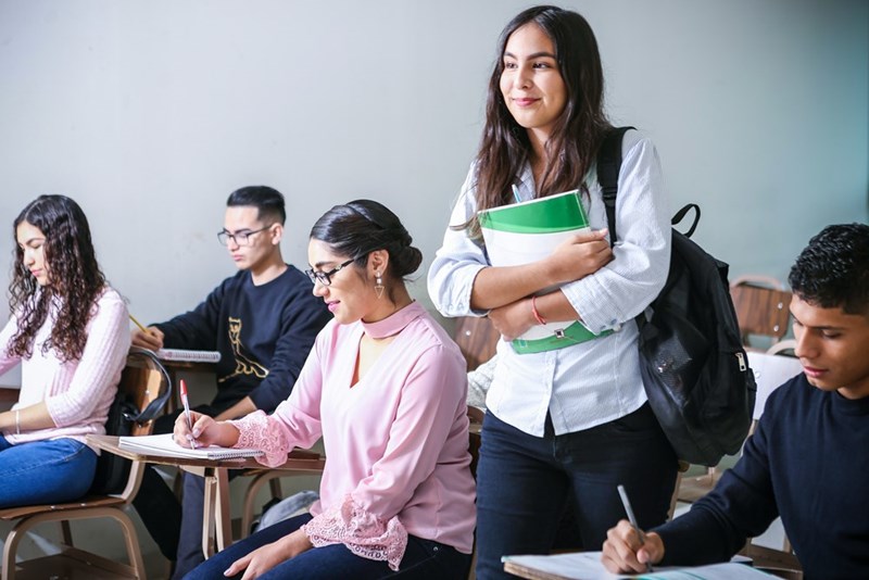 A college student with her books and her bag inside a classroom with other students. Representation.