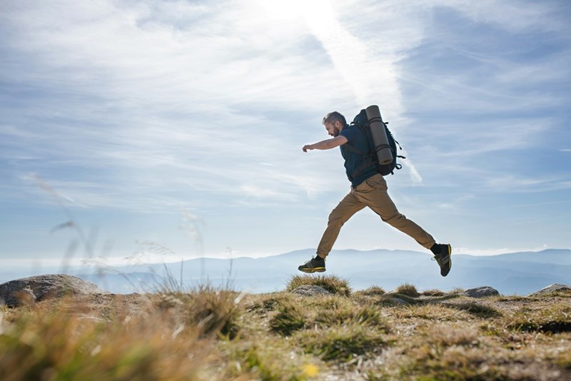 Man with a backpack jumps across rocks on a mountain ridge under a cloudy blue sky.