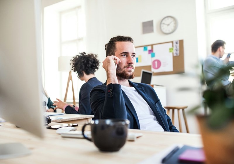 Man in a blazer sits at a desk in a bright office with coworkers blurred behind him.