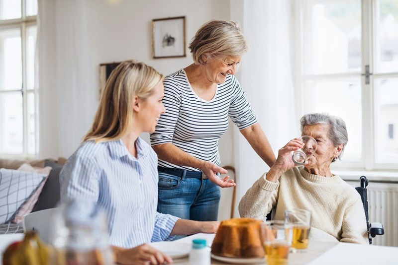 A live-in caregiver spends time with an elderly woman at home while another guest joins them at the table.