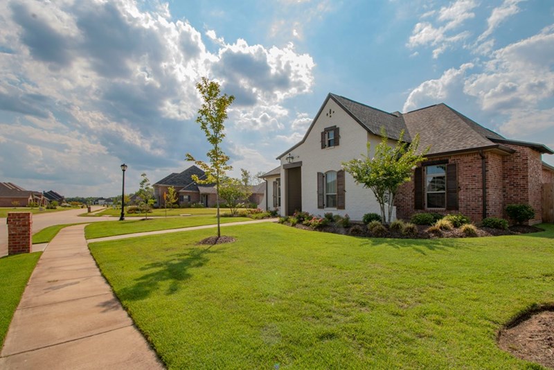 A large suburban home sits on a quiet residential street, surrounded by a landscaped lawn and neighboring properties.