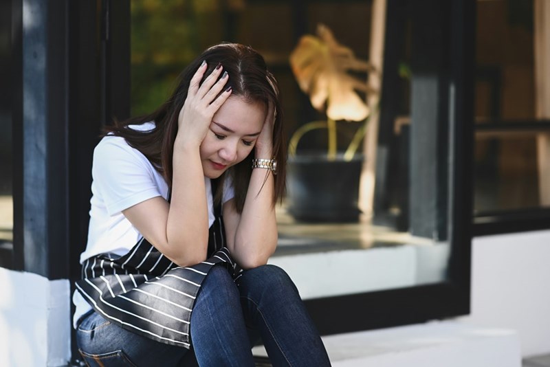 A restaurant worker sits outside with her head in her hands, appearing overwhelmed after what looks like a stressful shift.