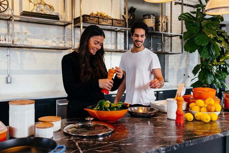 Two people prepare a meal together in a spacious kitchen, surrounded by cookware, fresh greens, and other ingredients.