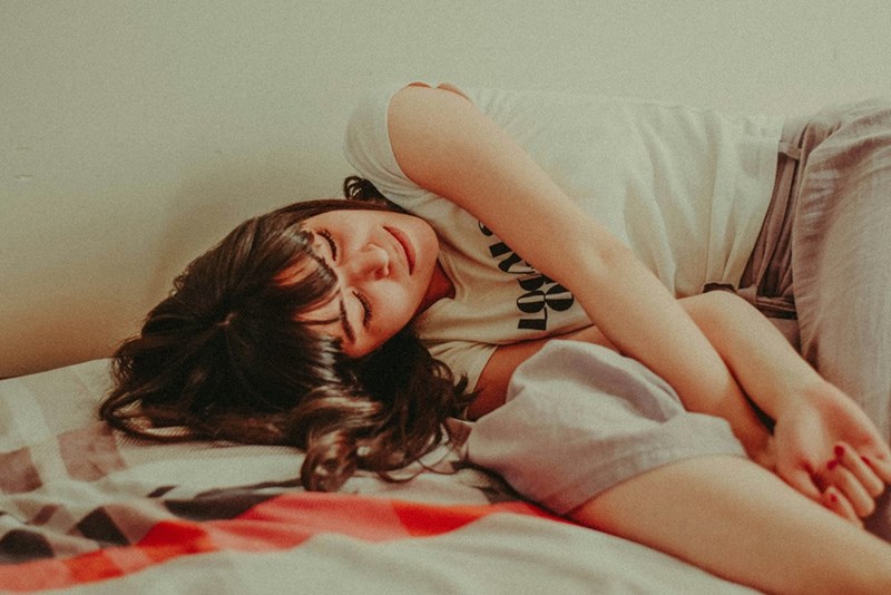 Young woman lies curled on a bed with eyes closed and a tense expression.
