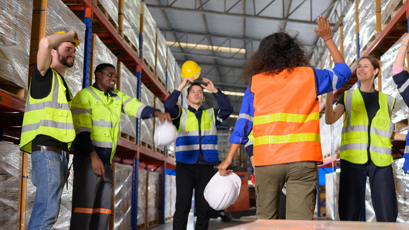 Warehouse workers in safety vests raise their hands between rows of shrink-wrapped pallets.