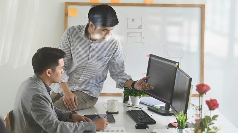 Two coworkers discussing a computer program at the office after a new hire developed a tool that transformed their daily workflow.