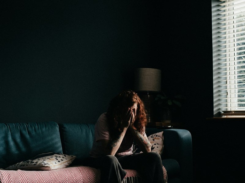 Red-haired woman sits on a couch in a dim room with her face covered by her hands.