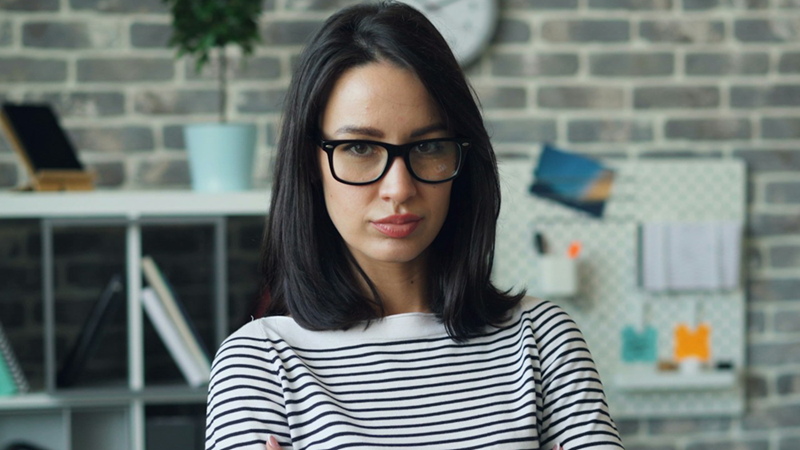 A representation of a female employee wearing glasses, standing in an office looking serious