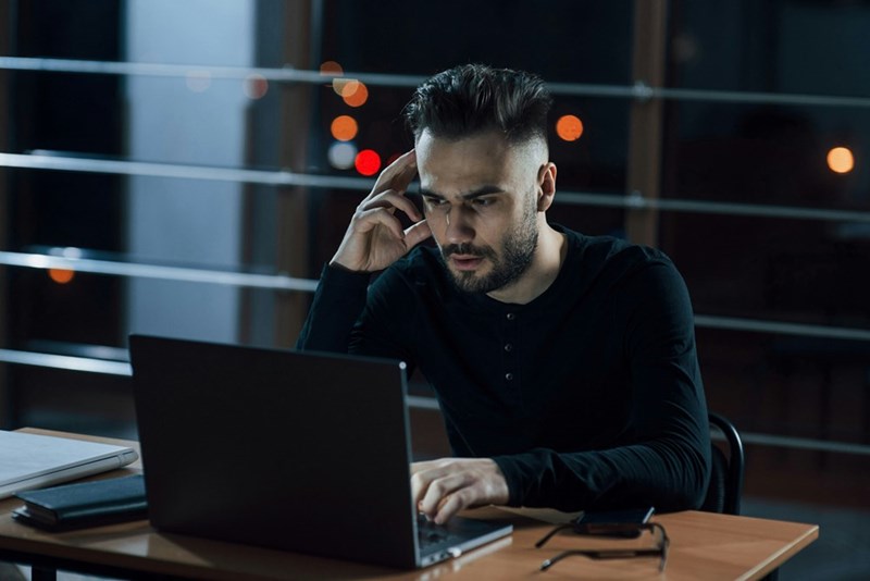Man sits at a laptop at night, holding a phone to his ear with a serious expression.