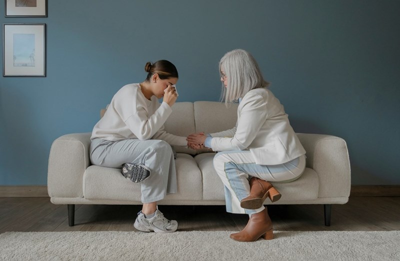 A mother and her adult daughter sit beside each other on the couch, crying