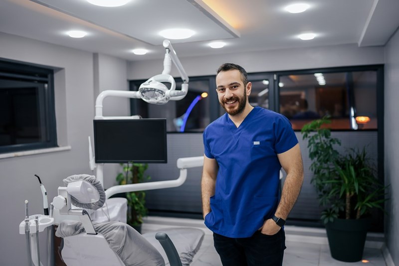 Dentist wearing blue scrubs stands inside a modern dental clinic beside a treatment chair and professional equipment.