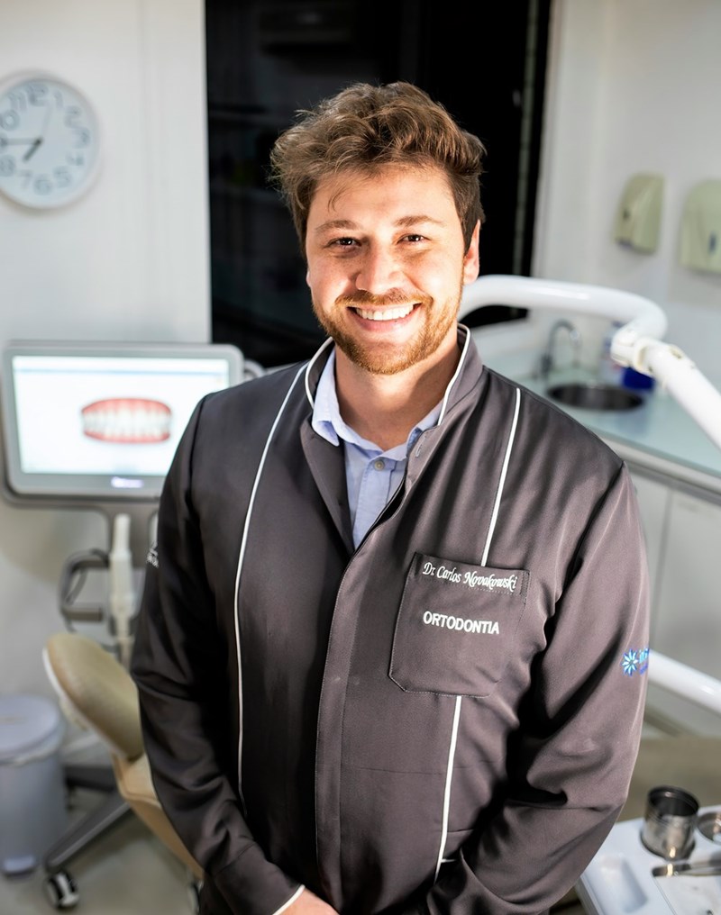 Smiling dental professional poses inside a dental office with treatment equipment and a digital teeth display in the background.