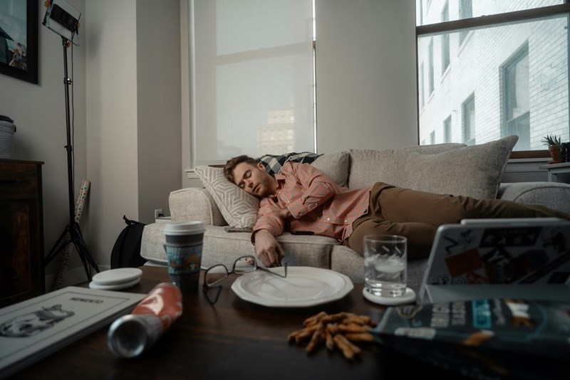 Man sleeps on a living room couch surrounded by dishes and clutter, depicting a relaxed moment at home.