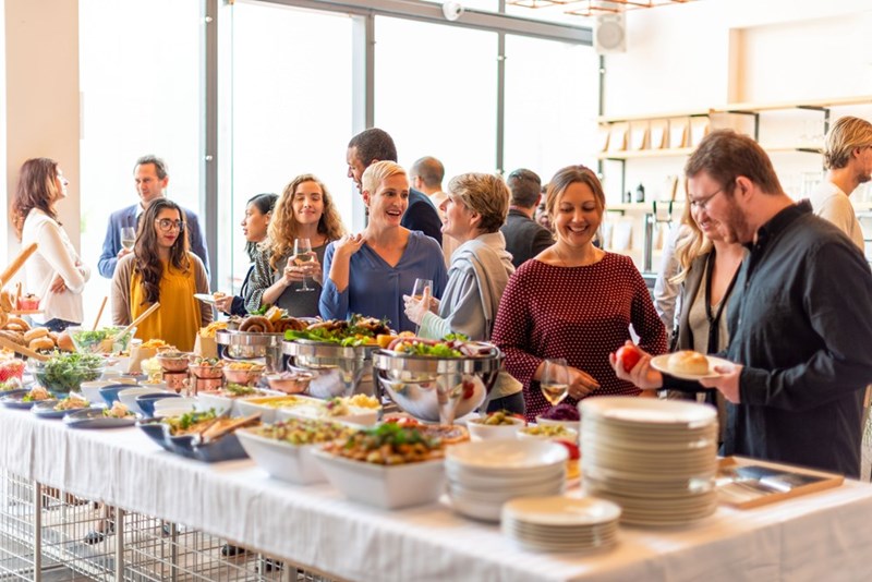 People gather around a buffet table with plates and drinks in a bright event space.
