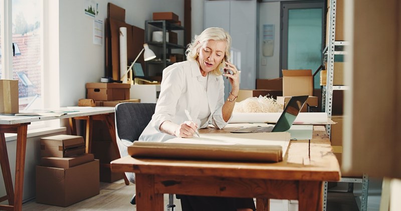 Blonde woman talks on a phone while writing at a desk surrounded by boxes and packing paper.