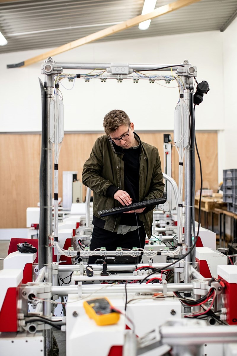 Man in glasses holds a keyboard and phone beside robotics equipment in a workshop.