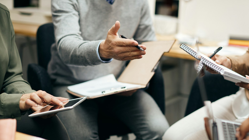 A close-up of colleagues sitting down in a circle while holding notebooks and tablets.