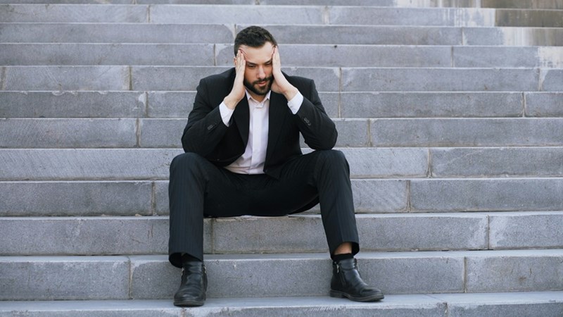 Man in a black suit sits on stone steps with both hands pressed to his head, looking stressed.
