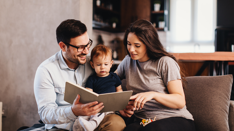 A couple and a young child sit on a couch looking together at an open book.