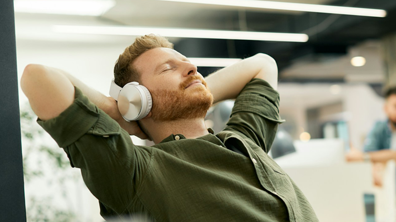 An employee wearing headphones sits in an office, leaning back with his eyes closed and his hands behind his head.