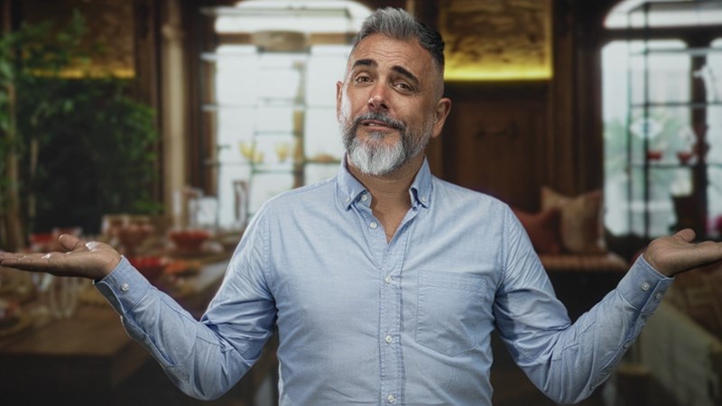Gray-bearded man in a blue shirt shrugs with both palms raised while standing indoors.