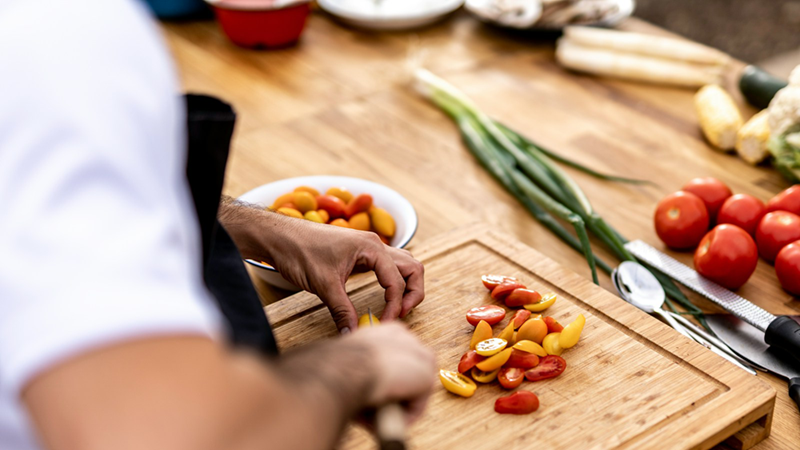 Close-up of a chef cutting tomatoes on a chopping board with ingredients and cutlery around him.