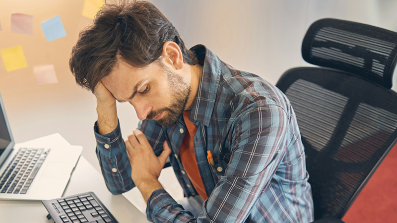 A male employee sitting at a desk with a computer and laptop runs his fingers through his hair.