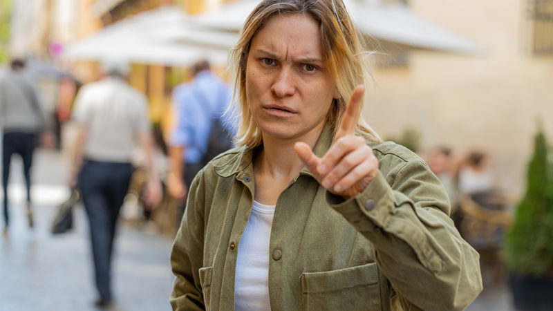 Woman in green jacket and white t shirt looking stern and confrontational points up like she's signaling "no"