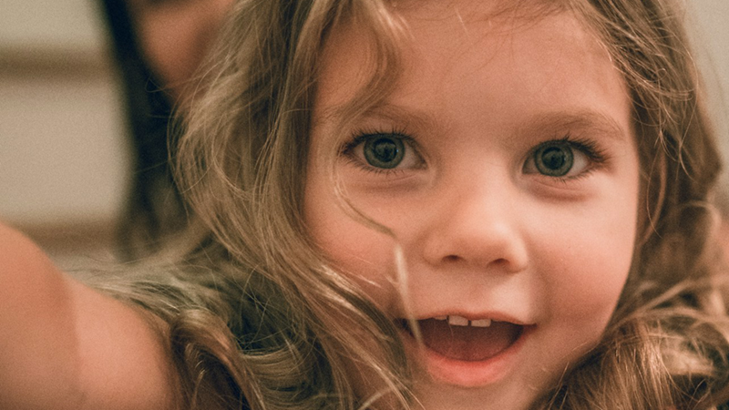 A close-up of a young girl with her arms outstretched, a woman in the unfocused background behind her.