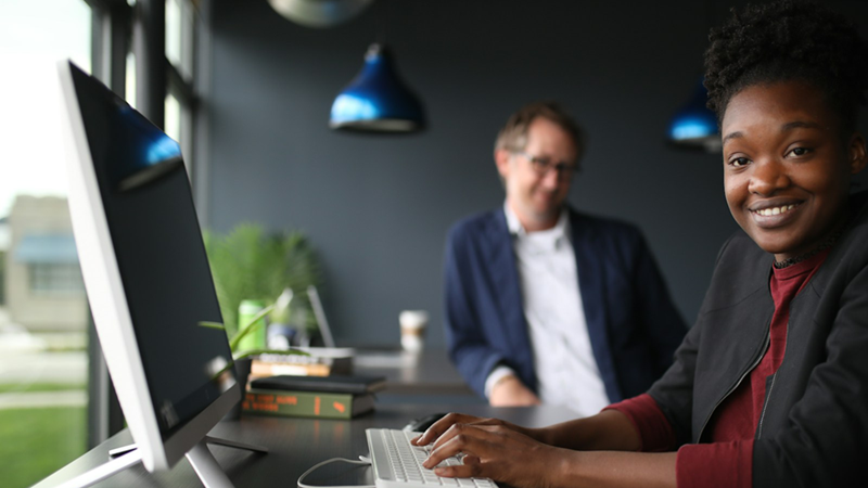 A female employee smiles while typing on a keyboard, a male coworker out of focus in the background behind her.