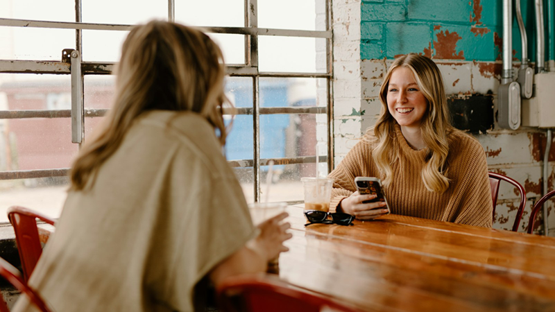 A woman sits across a table from another woman, smiling and holding her phone.