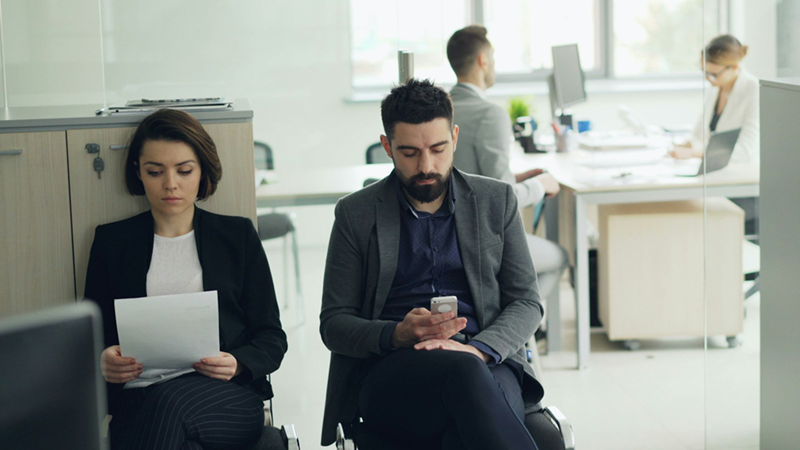 A woman and a man sit next to each other outside a glass-walled office with two people in it while she reads a piece of paper in her hand and he looks at his phone.