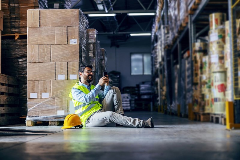 Young smiling employee sitting on the floor in warehouse, taking a break and using smart phone