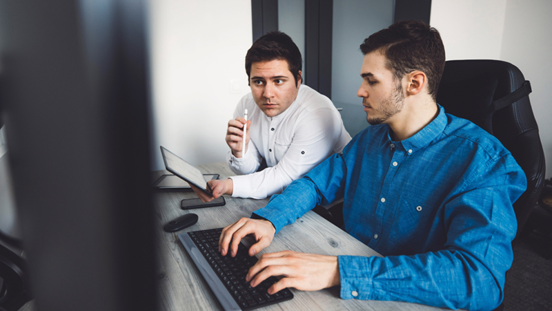 Two men sitting close to each other in a small office space, discussing something together