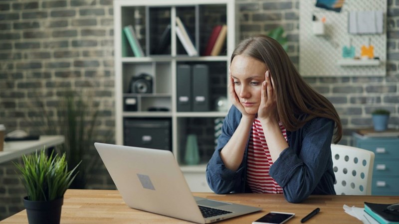 A woman sitting at a table with a laptop in front of her and looking burned out. Representative only.