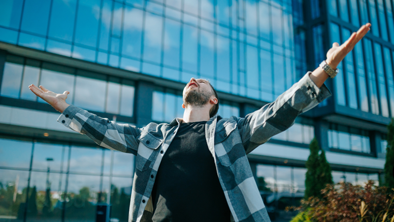 man looking up with arms open wide outside an office building.